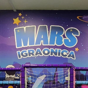 Logo "Mars igraonica" u velikim zutim slovima na ljubicasto-plavoj svemirskoj pozadini sa zvezdama i astronautom.