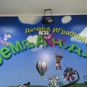 Zidni logo decije igraonice Zemlja cuda sa zelenim i zutim slovima i ilustracijama akrobata, balona i cvetova na plavom nebu.